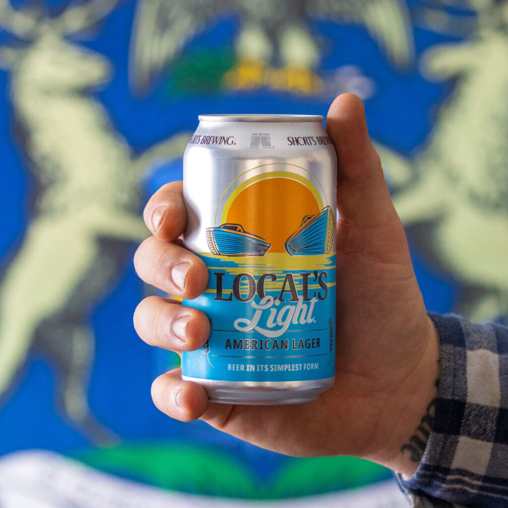 Photo of a hand holding a can of Local's Light lager in front of the Michigan Flag