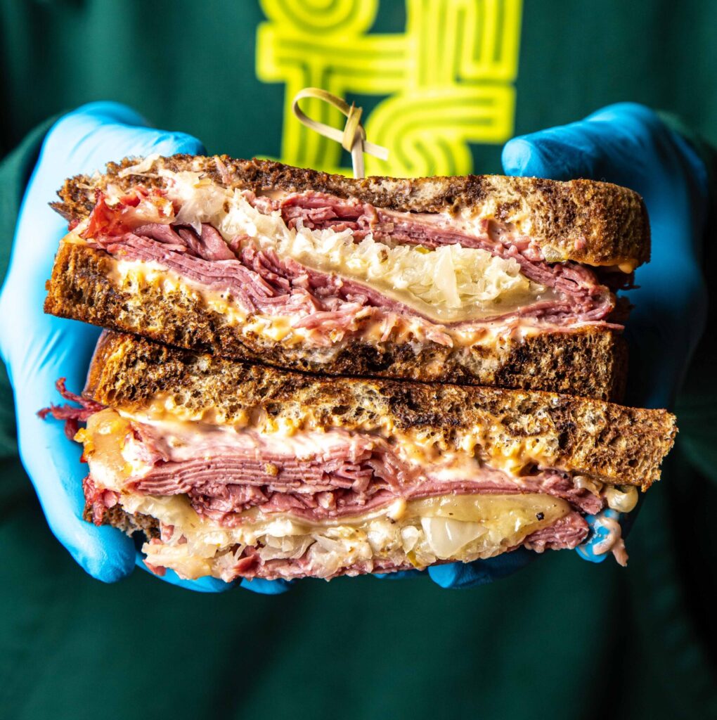 Photo of hands holding a reuben sandwich (we call The Rift) sliced in half